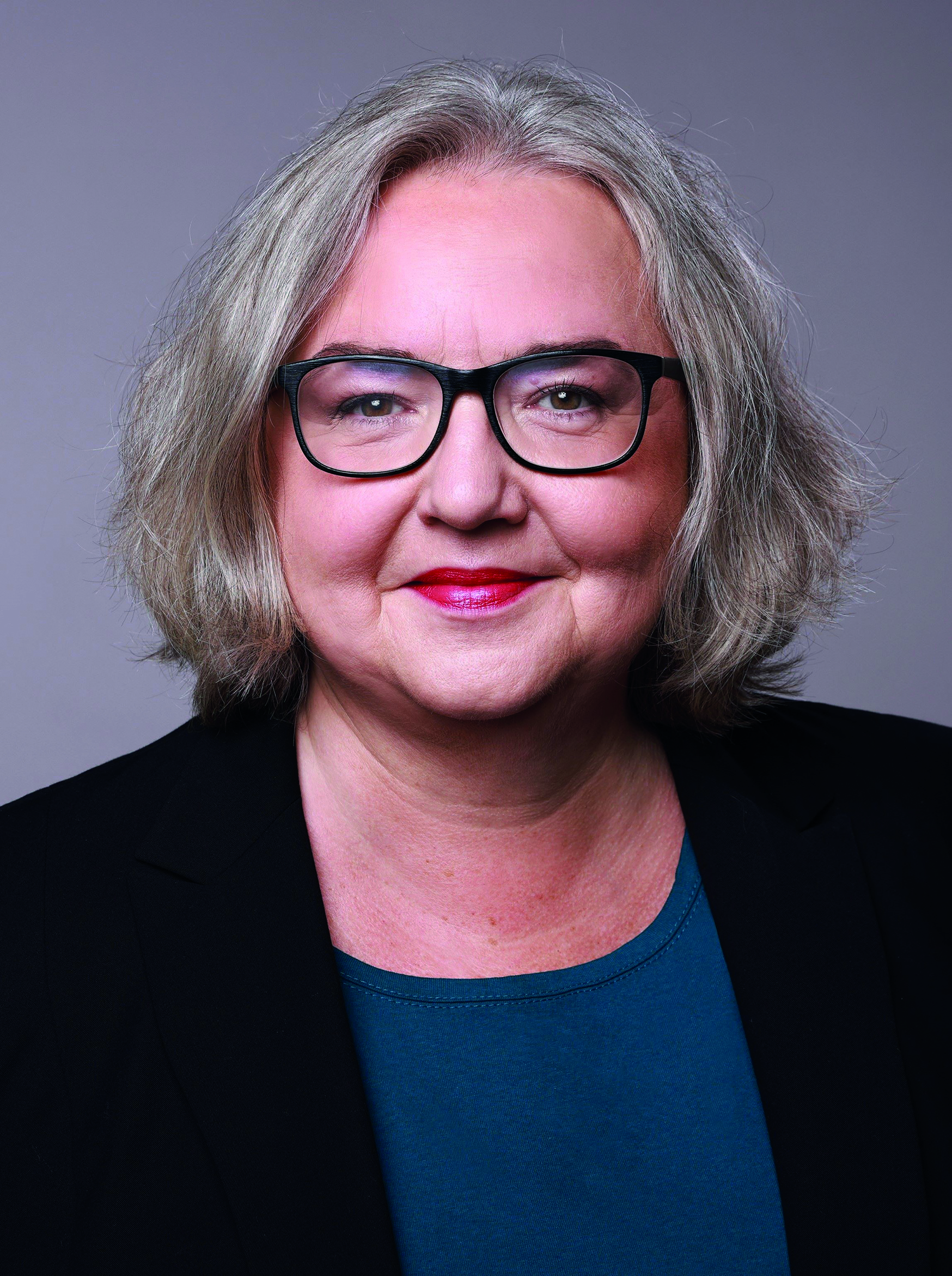 Grauharige Frau mit Brille und rotem Lippenstift vor grauem Hintergrund.