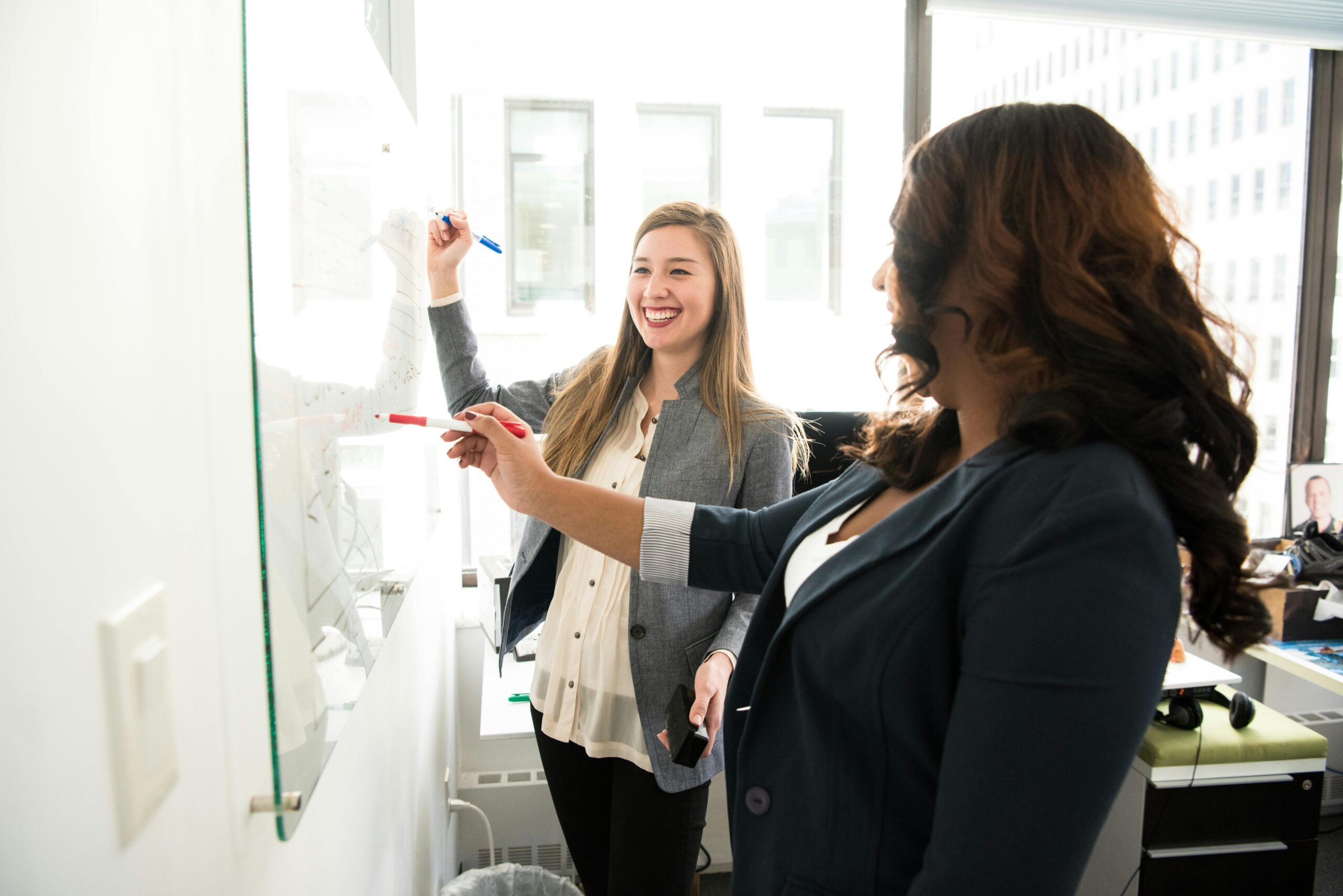 Zwei Frauen schreiben lächelnd mit Stiften auf ein Whiteboard in einem Büro.