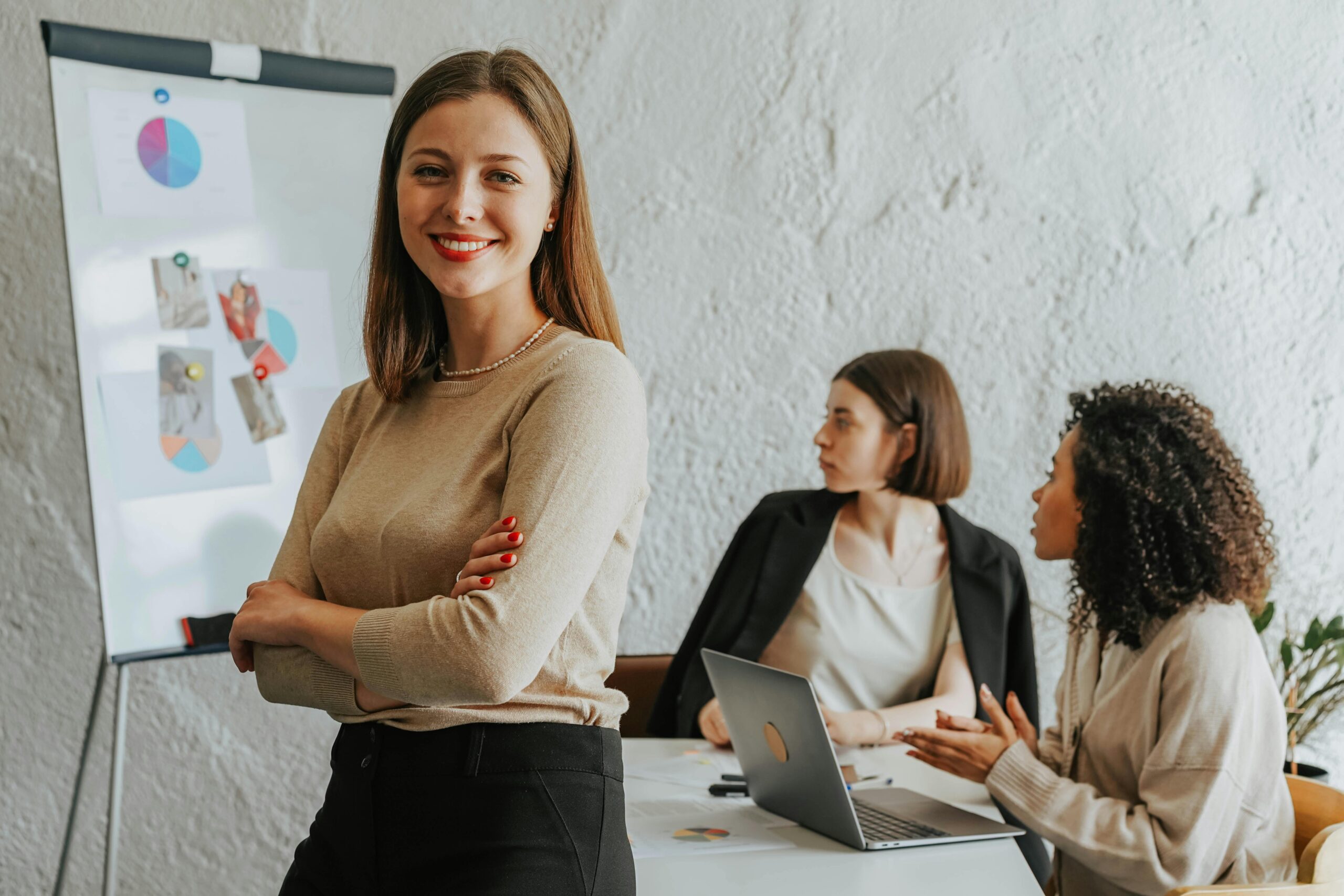 Frau vor Flipchart lächelt, zwei Frauen am Tisch mit Laptop im Gespräch.