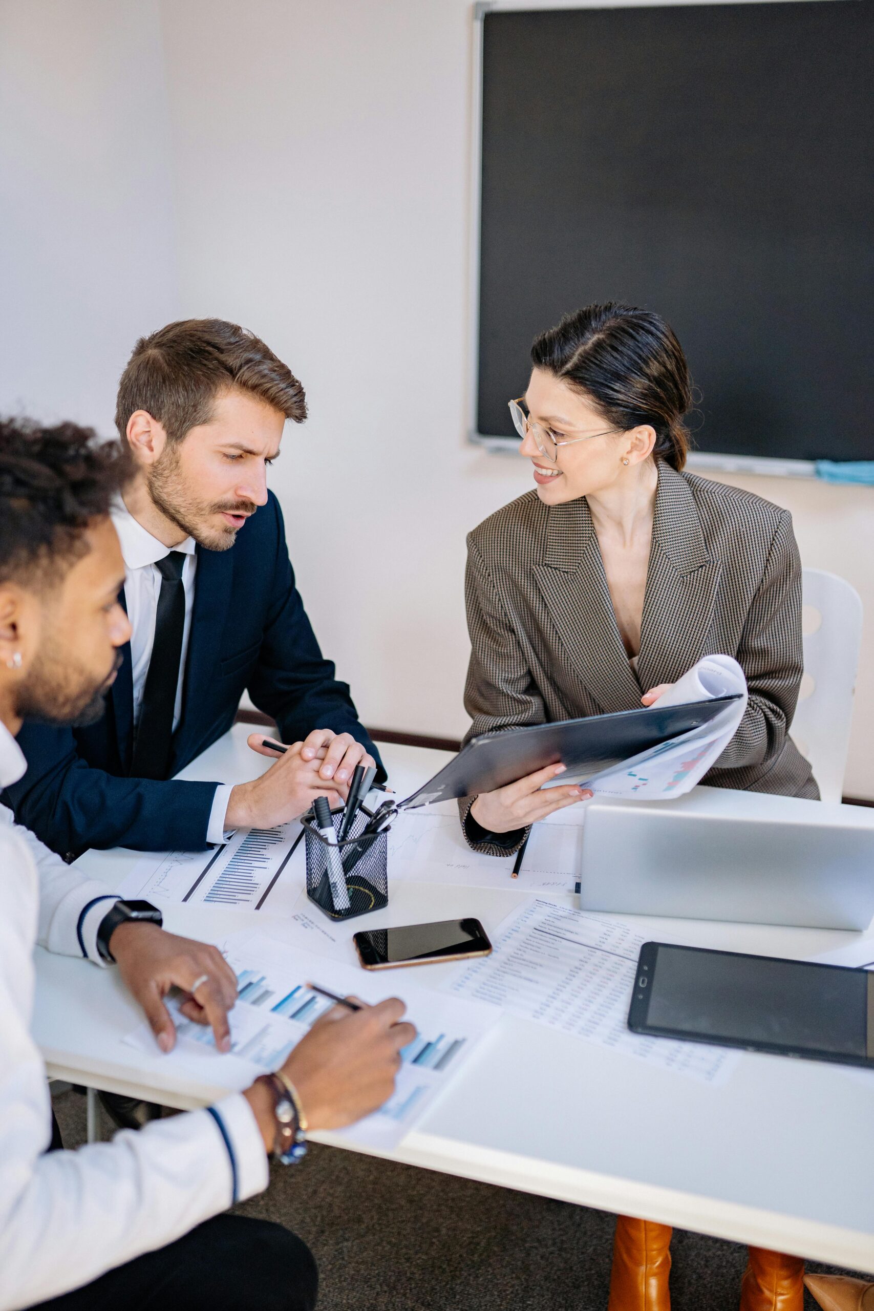 Drei Personen in formeller Kleidung diskutieren an einem Tisch mit Dokumenten.