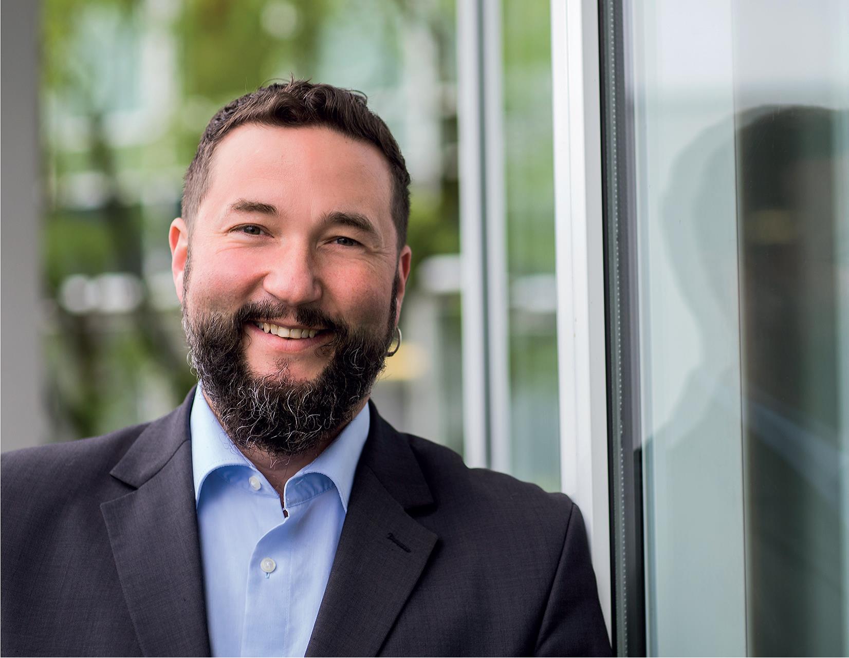 Eine lächelnde Person in Anzug steht neben einem Fenster.