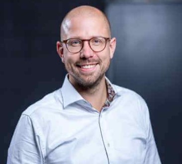 Eine lächelnde Person mit Brille und weißem Hemd vor unscharfem Hintergrund.