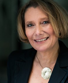 Eine lächelnde Frau mit kurzem, blonden Haar in dunkler Kleidung vor unscharfem Hintergrund.