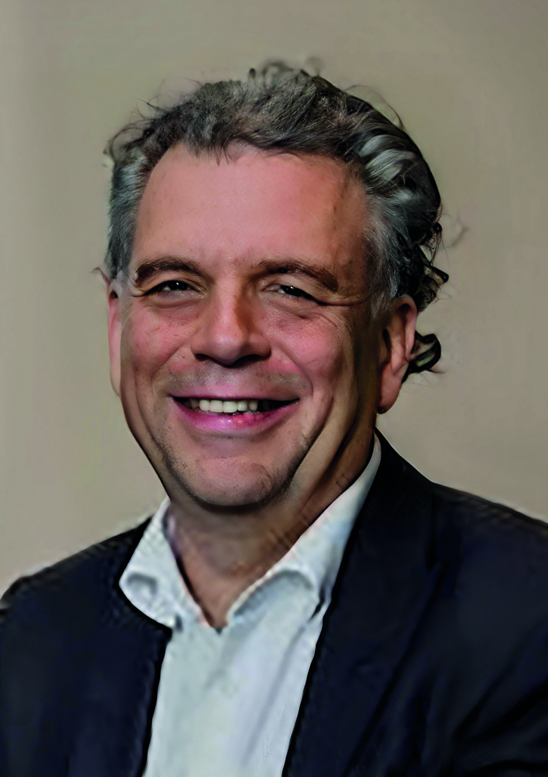 Mann mit grauem Haar in Anzug lächelt vor neutralem Hintergrund.