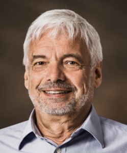 Mann mit grauem Haar und Bart in hellblauem Hemd vor braunem Hintergrund.