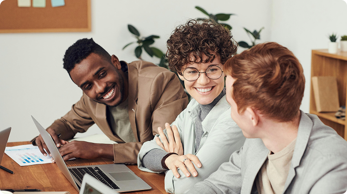 Drei Personen sitzen lächelnd an einem Tisch mit Laptops und Dokumenten.