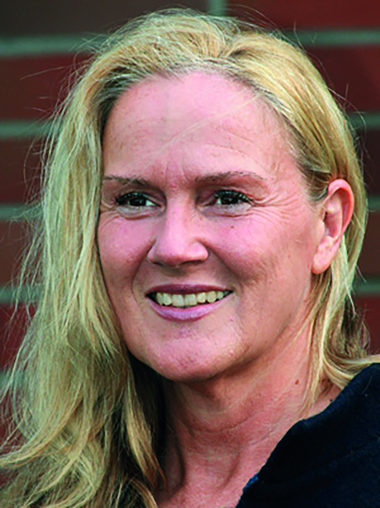 Eine lächelnde Person mit langen blonden Haaren vor einer roten Backsteinmauer.