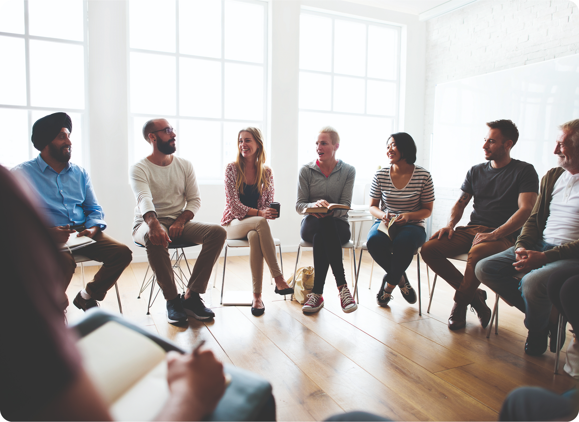 Menschen sitzen in einem hellen Raum im Stuhlkreis und unterhalten sich lächelnd.