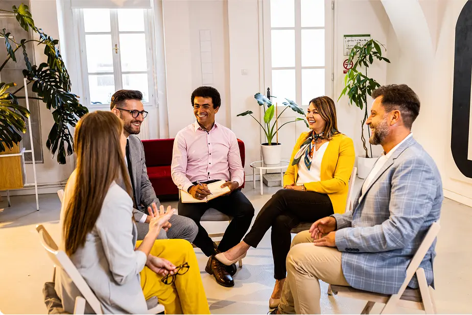 Sechs Personen sitzen lächelnd in einem Kreis in einem hellen Raum mit Pflanzen.