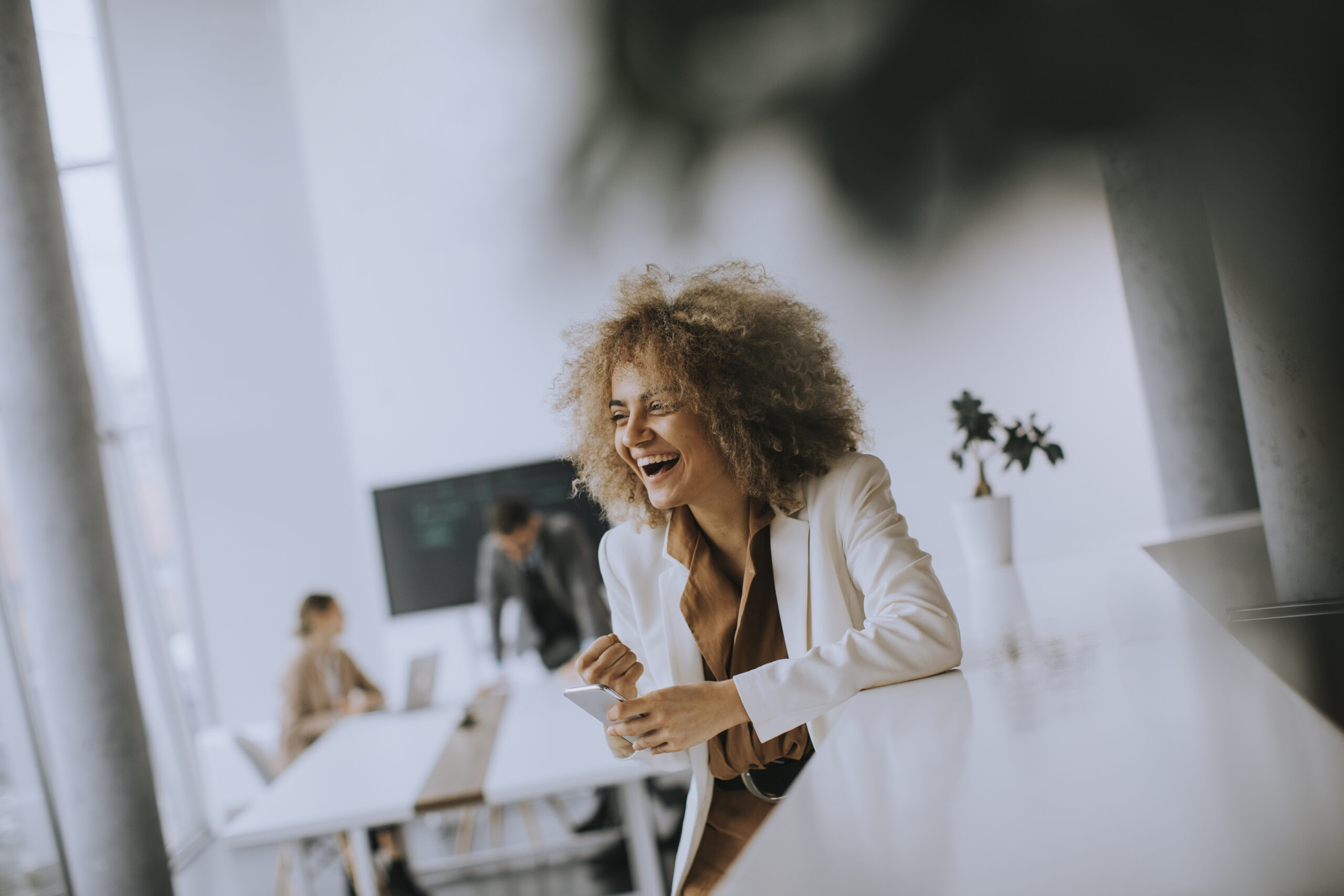 Person in weißem Blazer lacht, im Hintergrund Personen an einem Tisch und ein Bildschirm.